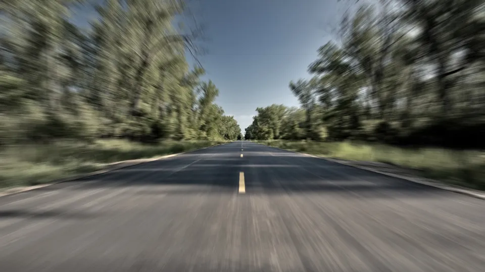 A blurred asphalt road stretches into the distance, flanked by trees on either side under a clear blue sky.