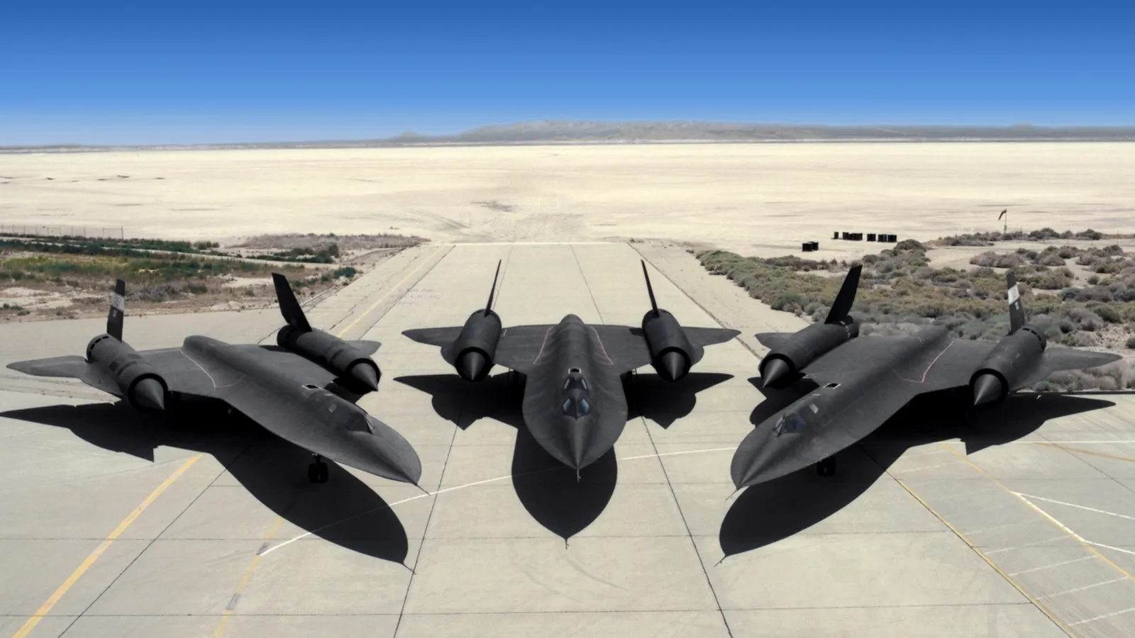 Three black SR-71 Blackbird spy planes are parked on a concrete tarmac in a desert landscape.