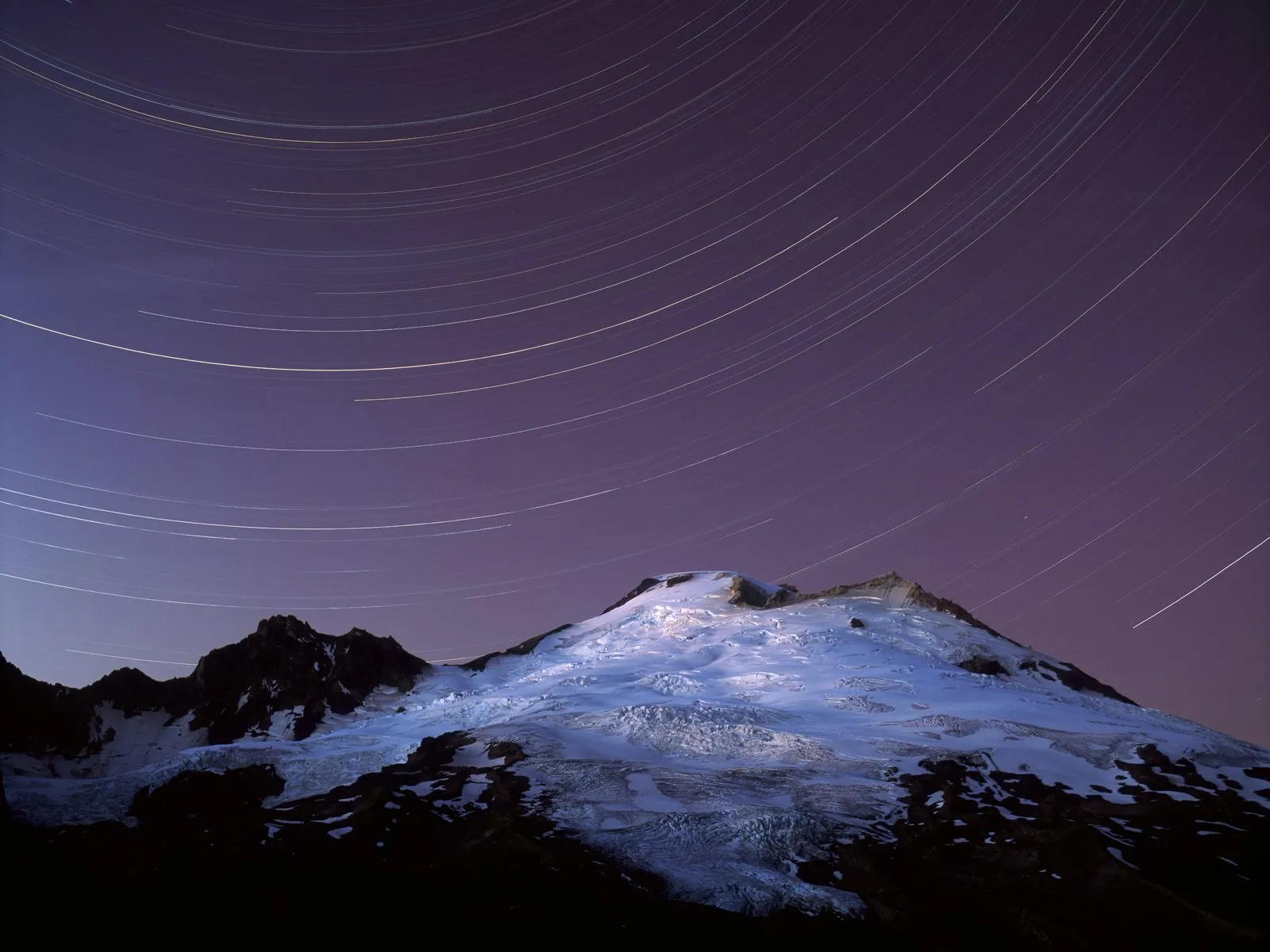Star trails arc across a deep purple night sky above a snow-covered mountain.