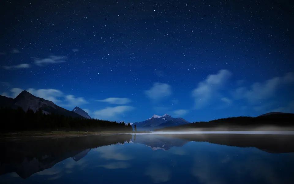 A clear night sky filled with stars above a calm lake reflecting the mountains and sky.