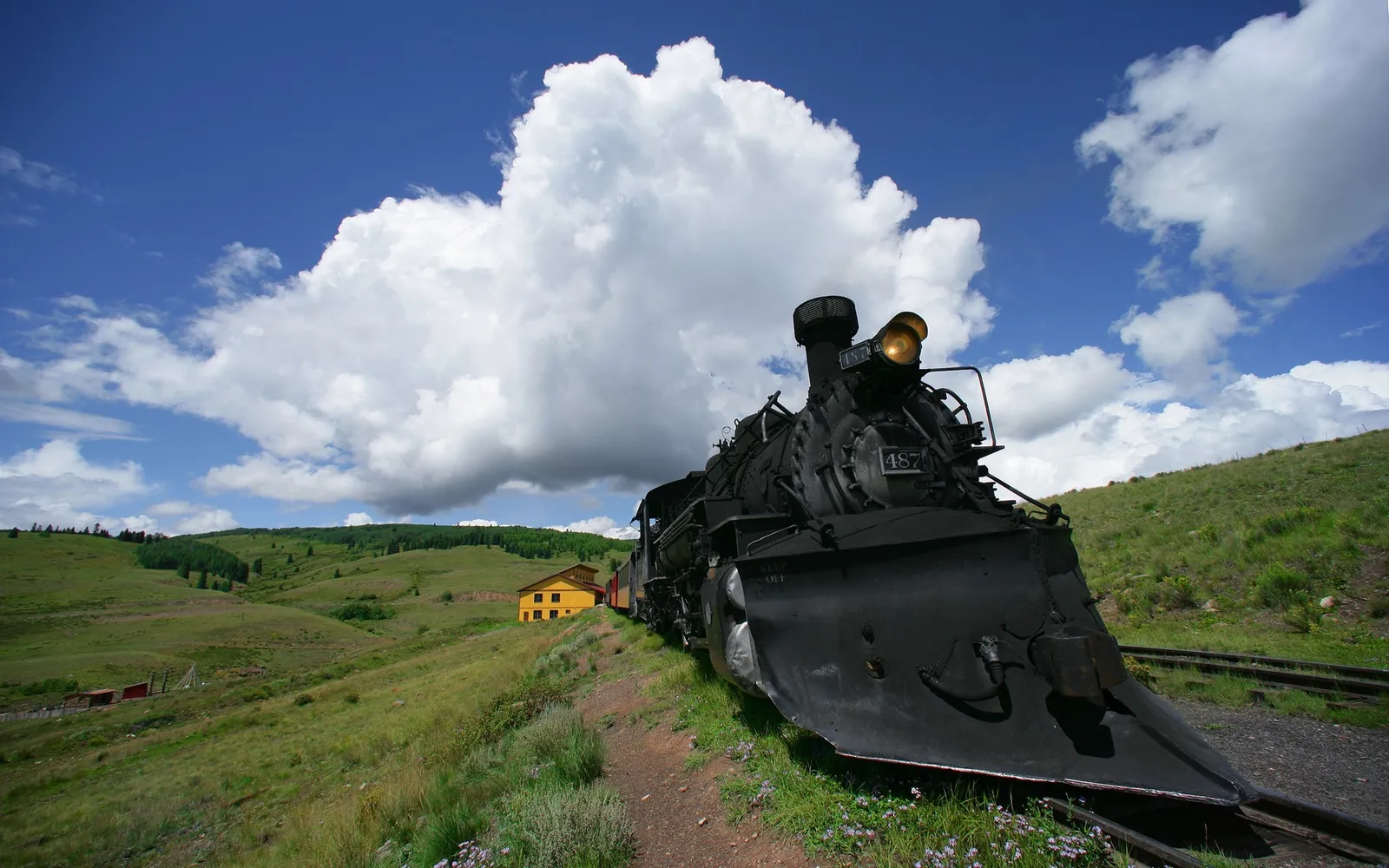 A black steam train is parked on tracks next to a grassy hill with a yellow building in the background under a blue sky with clouds.