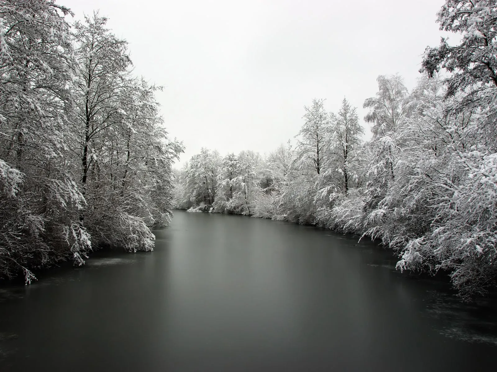A wide, dark, frozen river is surrounded by trees heavily coated in snow under an overcast sky.