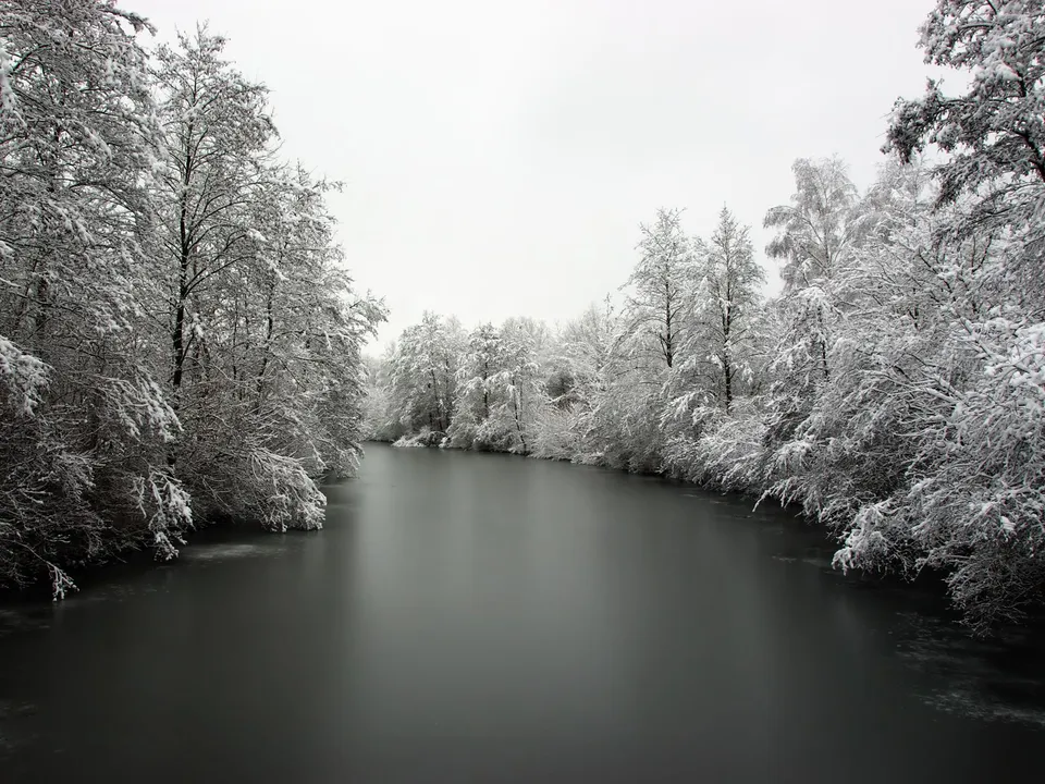 A wide, dark, frozen river is surrounded by trees heavily coated in snow under an overcast sky.