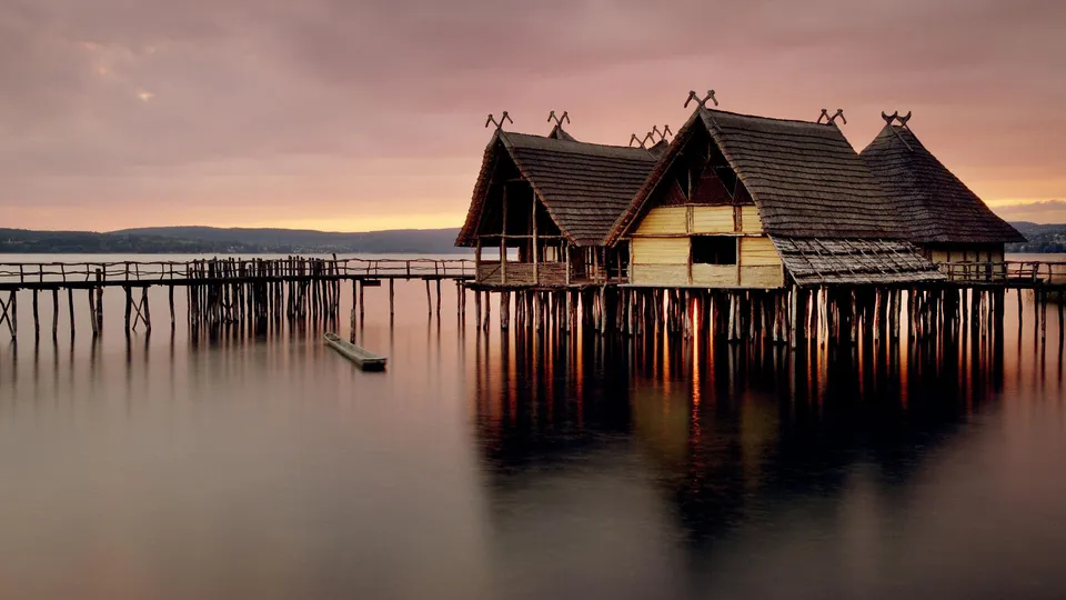Wooden stilt houses with thatched roofs stand over calm water, reflecting the soft colors of a sunset sky.