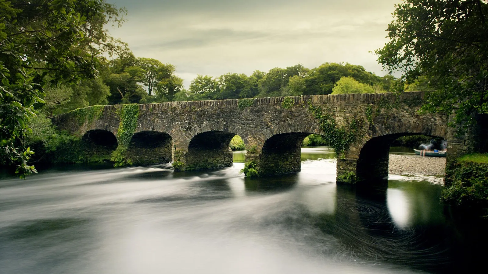 A long exposure shot of a stone bridge with ivy over a flowing river, with trees in the background.