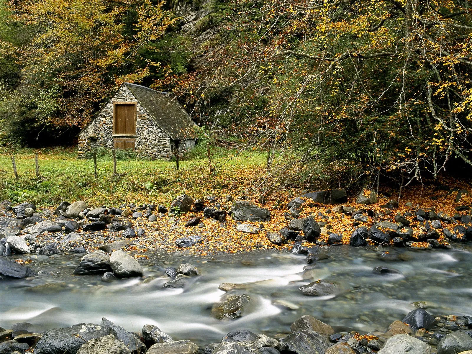 A small stone cabin with a wooden door sits beside a flowing stream in an autumn forest.