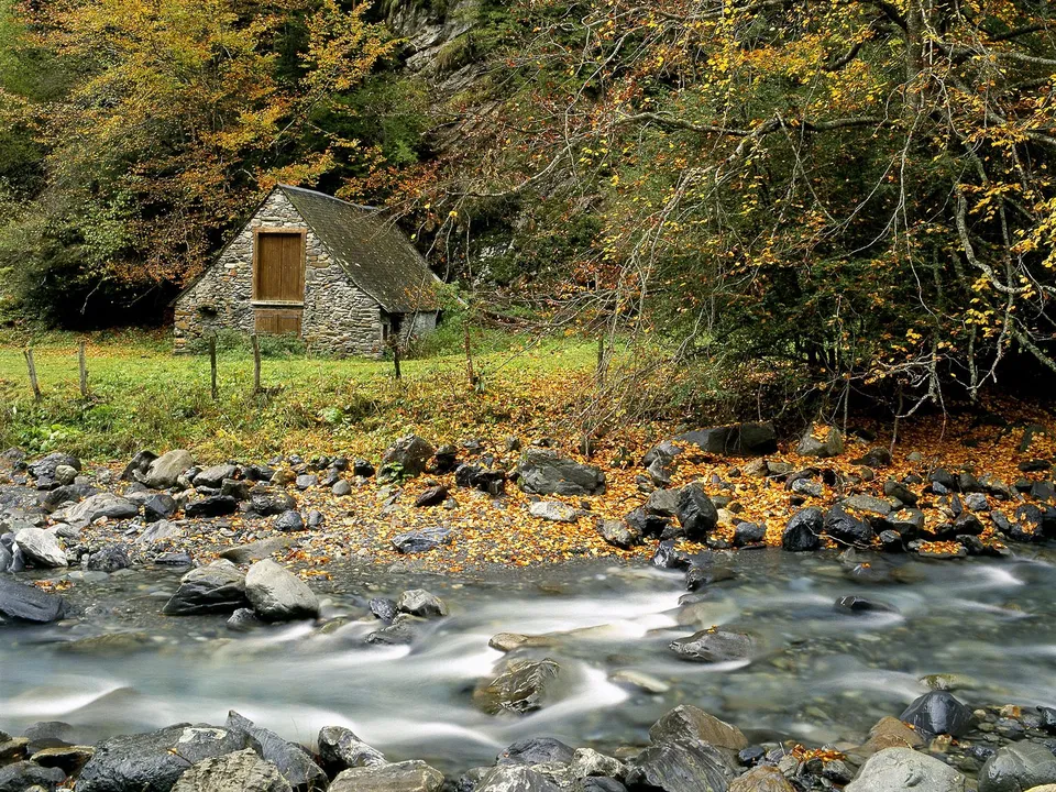A small stone cabin with a wooden door sits beside a flowing stream in an autumn forest.