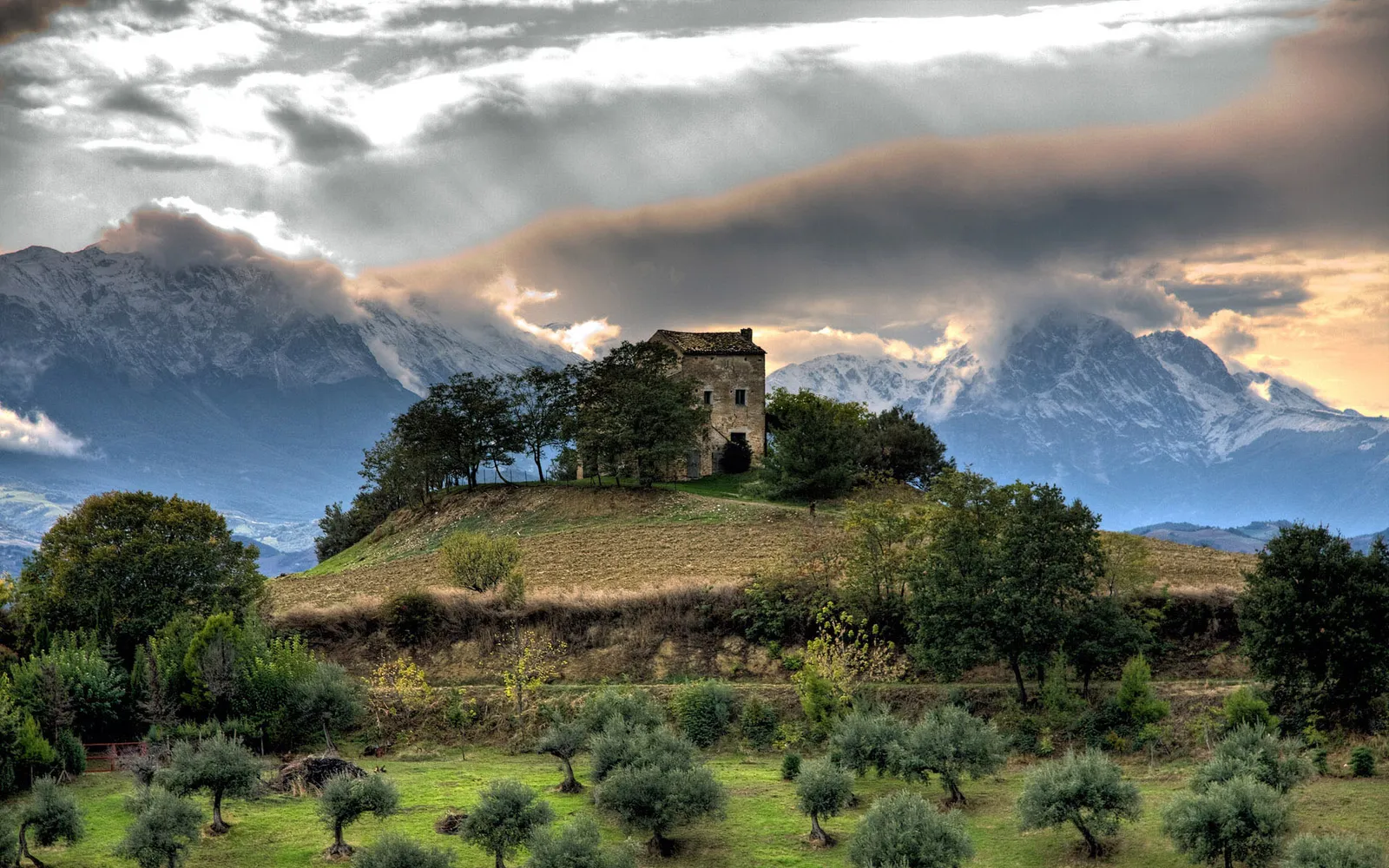 A stone house sits on a grassy hill with snow-capped mountains in the background under a cloudy sky.