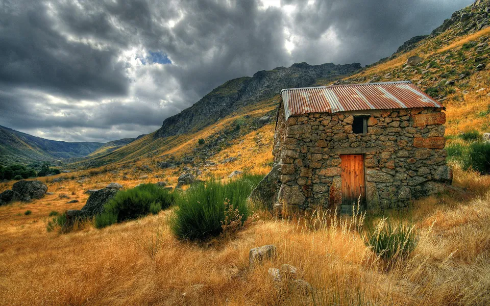 A weathered stone hut with a corrugated metal roof sits on a dry, grassy hillside under a dramatic, cloudy sky.
