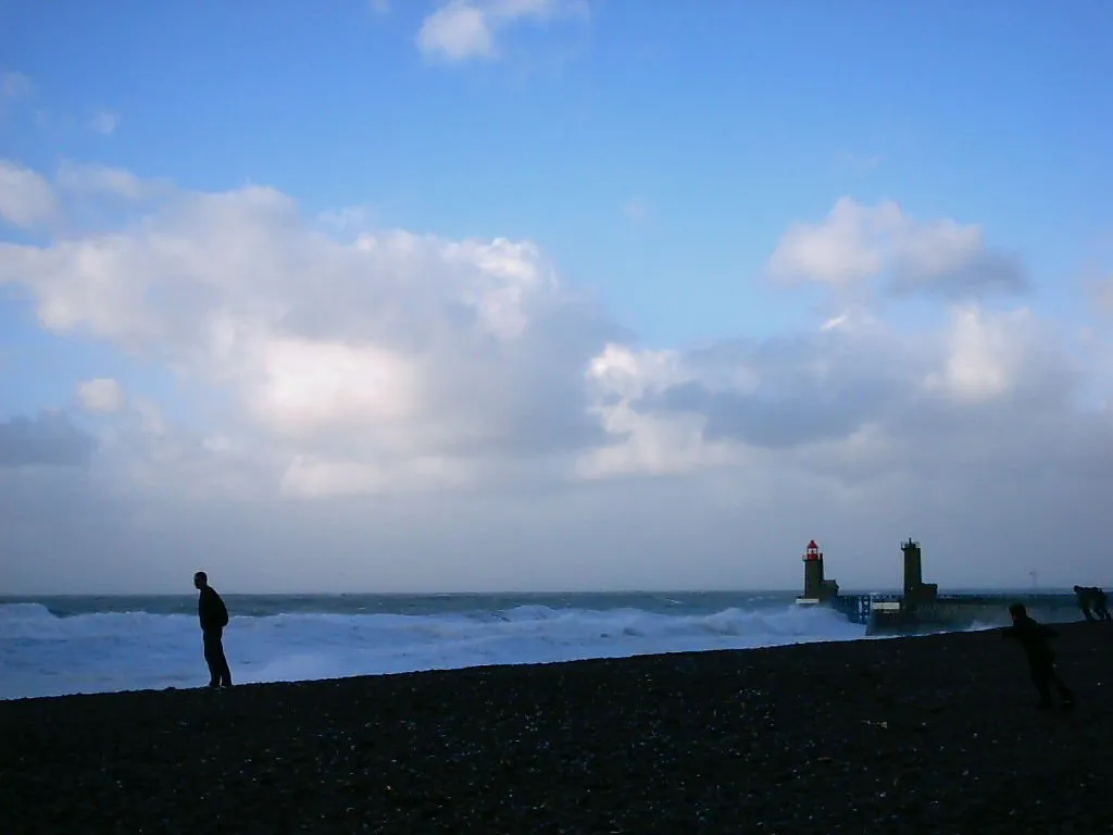 A person stands on a dark, pebbly beach watching large waves crash near a lighthouse in the distance.