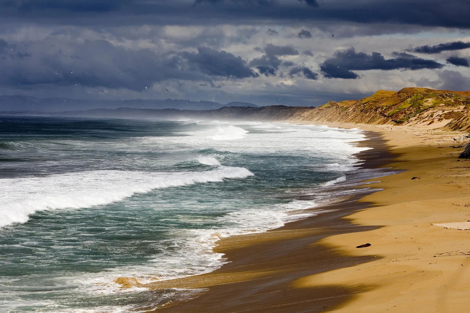 A wide view of a sandy beach with powerful waves crashing ashore under a dark, stormy sky.