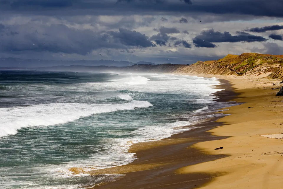 A wide view of a sandy beach with powerful waves crashing ashore under a dark, stormy sky.