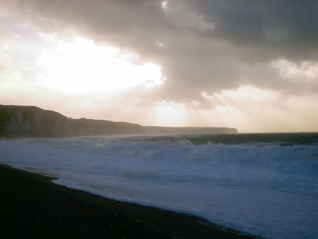 Waves crash onto a dark pebble beach with dramatic clouds and sunbeams overhead.
