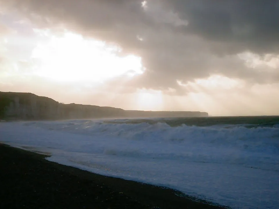 Waves crash onto a dark pebble beach with dramatic clouds and sunbeams overhead.