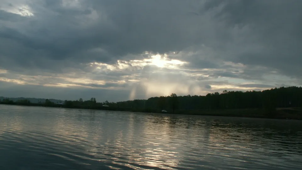 Sunlight streams through dark clouds onto a rippling lake, with a tree-lined shore in the background.