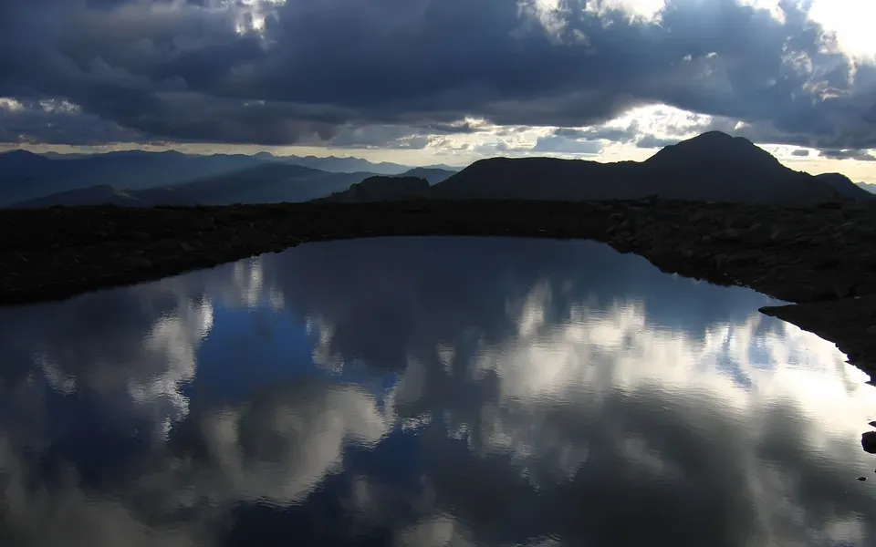 A dark mountain lake reflects dramatic, stormy clouds and silhouetted mountains.