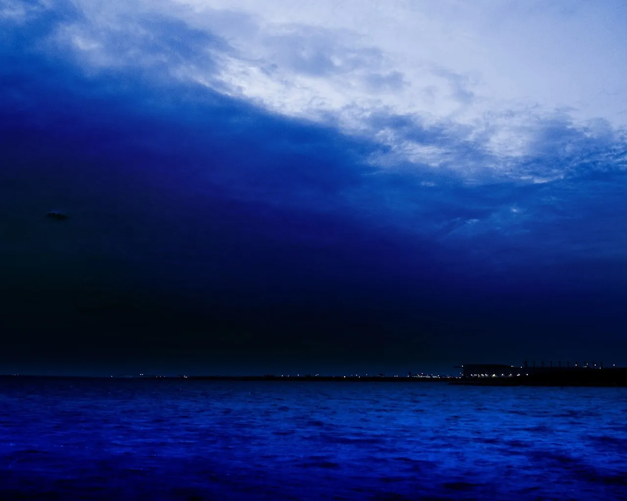 A dark, stormy sky with deep blue clouds looms over a calm ocean at dusk, with distant lights on the horizon.