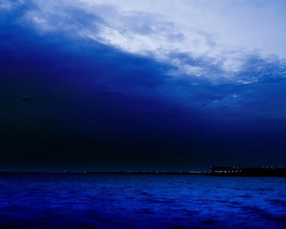 A dark, stormy sky with deep blue clouds looms over a calm ocean at dusk, with distant lights on the horizon.