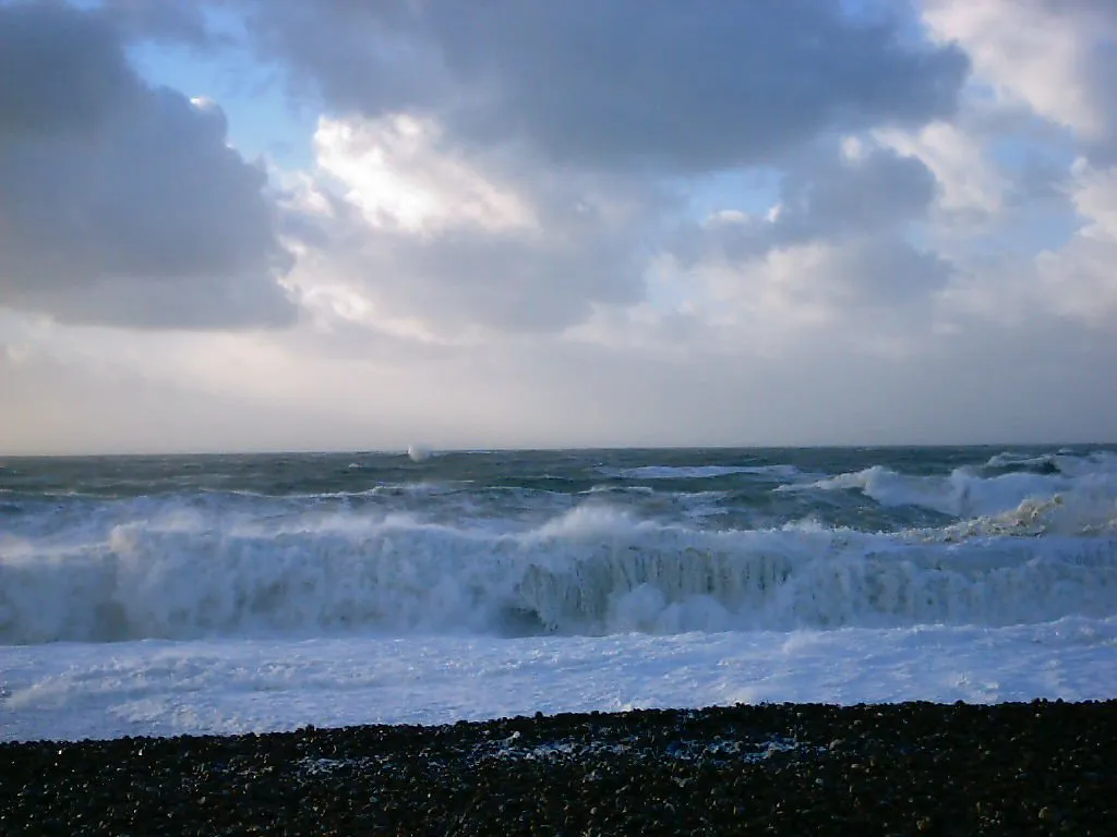Powerful waves crash onto a dark pebble beach under a dramatic, cloudy sky.