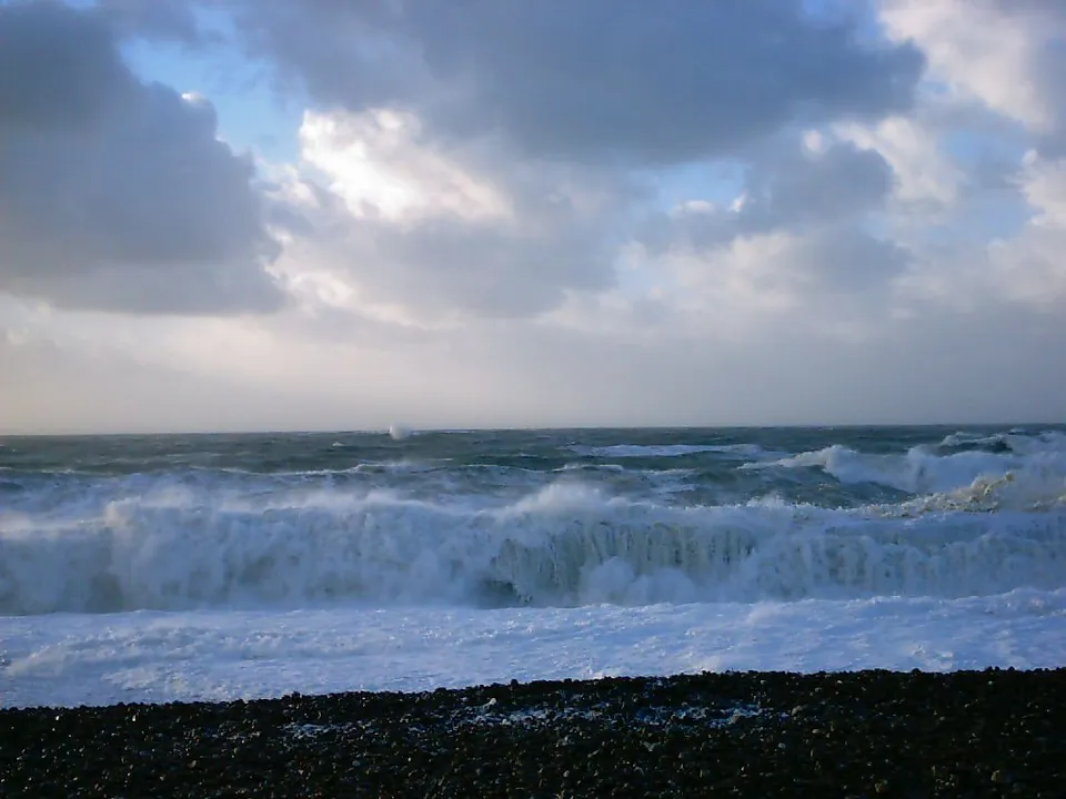Powerful waves crash onto a dark pebble beach under a dramatic, cloudy sky.