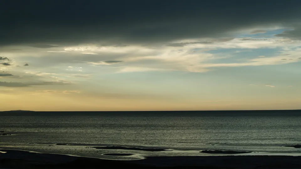 Dark storm clouds gather over a tranquil sea with a faint horizon line and a distant island.
