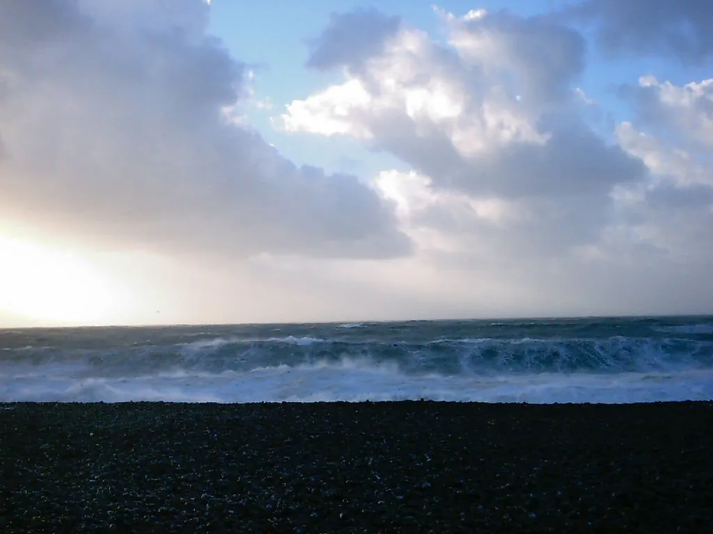 A dark pebble beach in the foreground with a rough sea and choppy waves under a dramatic sky with clouds.