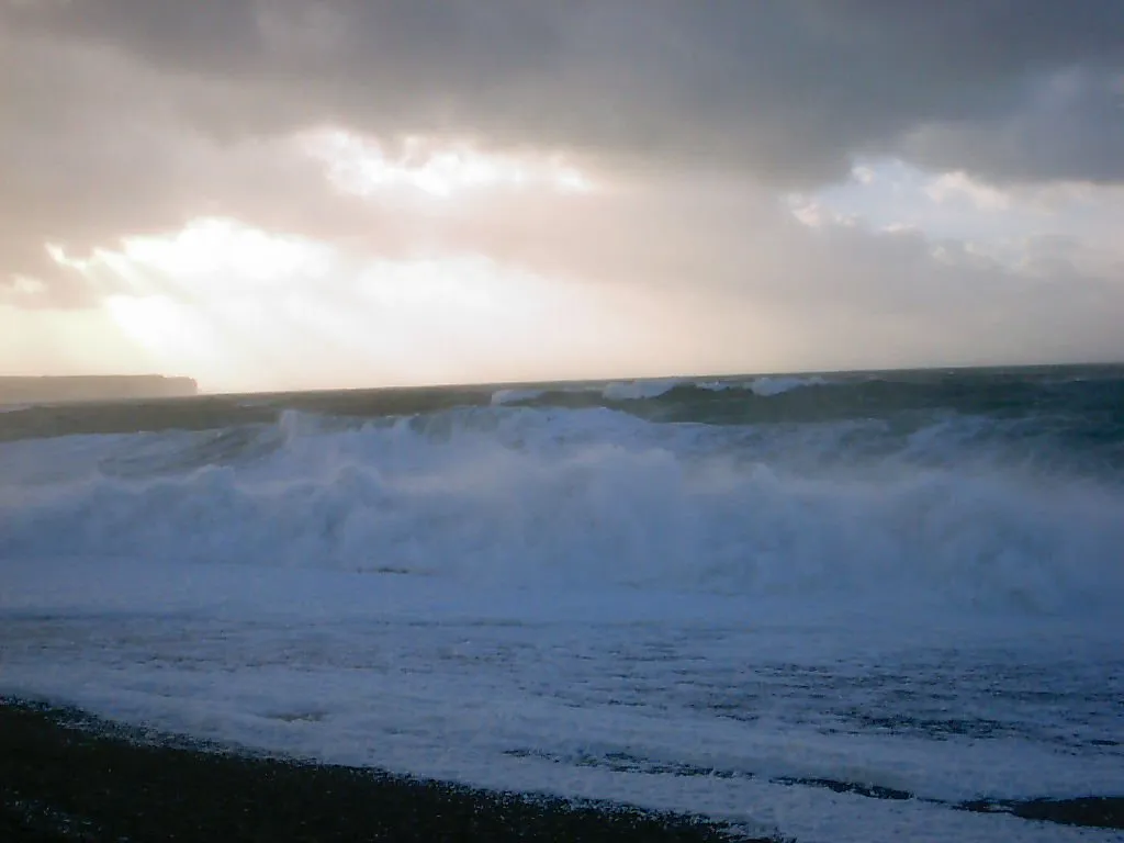 Waves crash on a dark pebble beach under a dramatic sky with rays of light breaking through dark clouds.