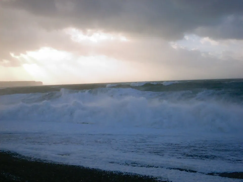 Waves crash on a dark pebble beach under a dramatic sky with rays of light breaking through dark clouds.