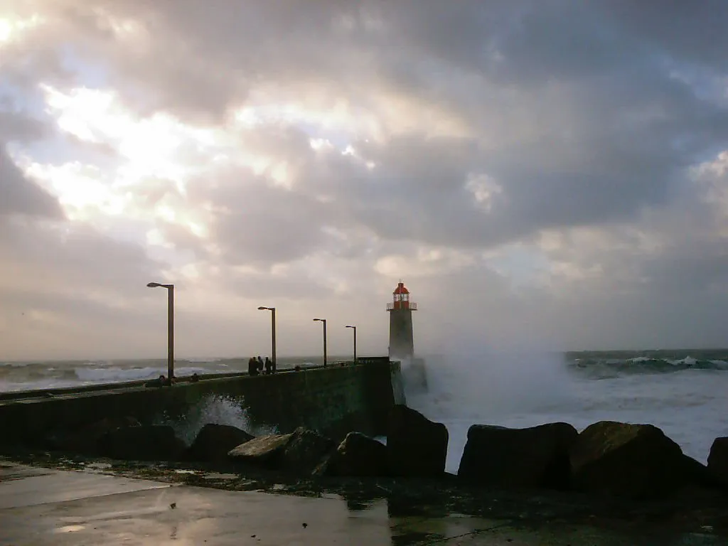 A lighthouse with a red top stands against stormy seas crashing on a pier.