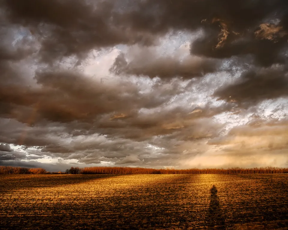 A dramatic sky with dark, heavy clouds and shafts of golden light over a dry, harvested field.