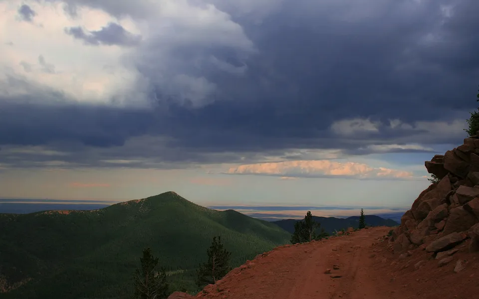 A dirt road winds towards green mountains under a dark, stormy sky.