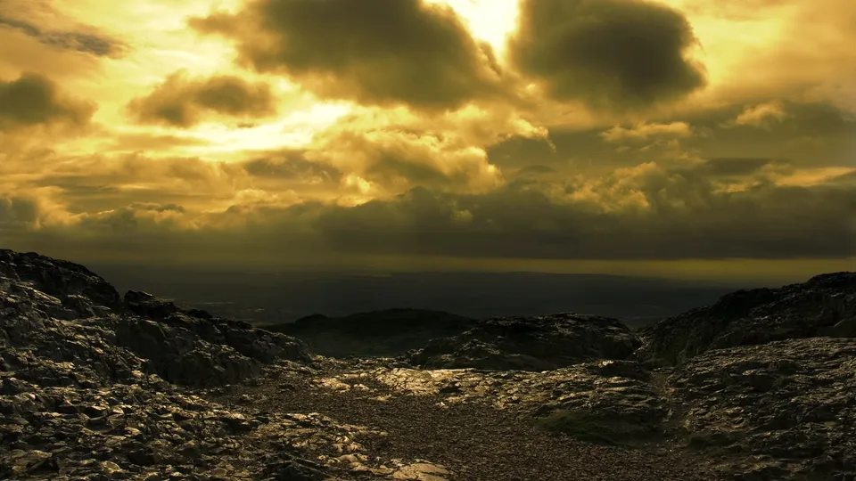 A dramatic, cloudy sky with golden light filters through the clouds over a rocky mountain landscape.