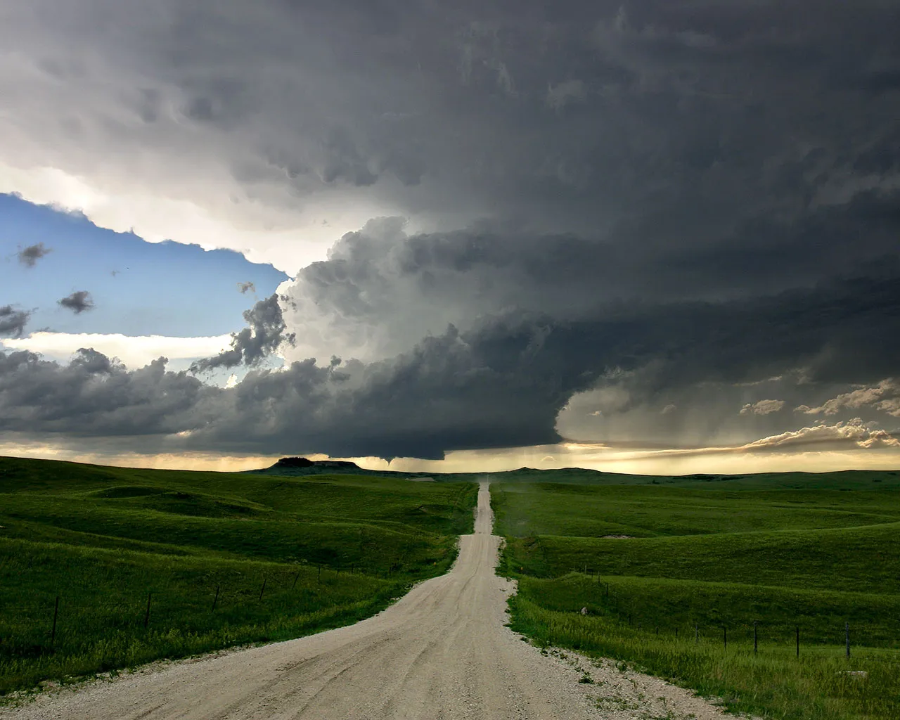 A dirt road leads through green rolling hills towards a dramatic, dark storm cloud formation.