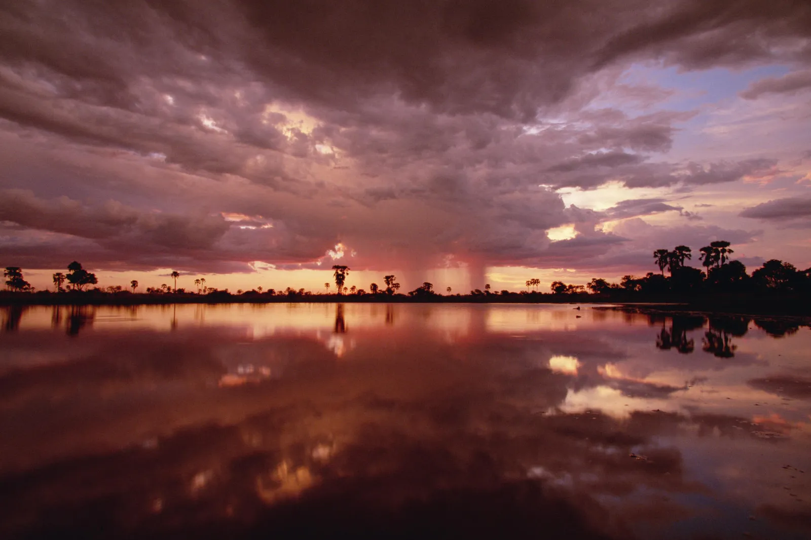 A dramatic sunset with heavy clouds reflected in a calm body of water, with silhouetted trees on the horizon.