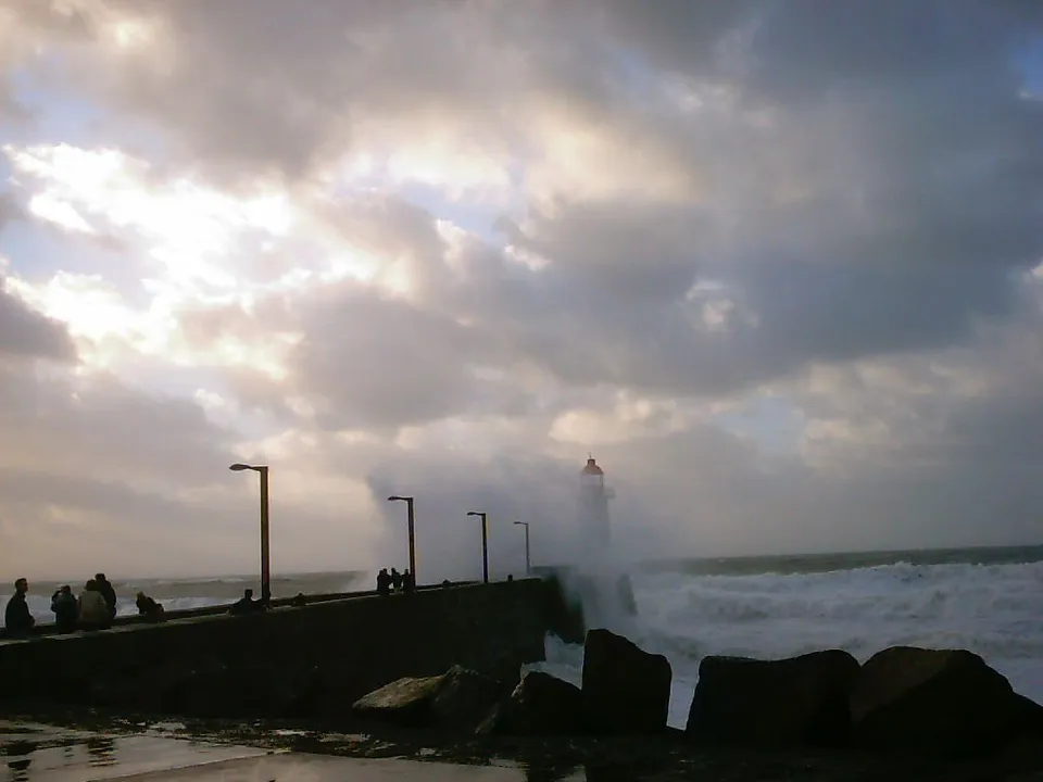 Stormy waves crash against a stone pier with a lighthouse in the distance.