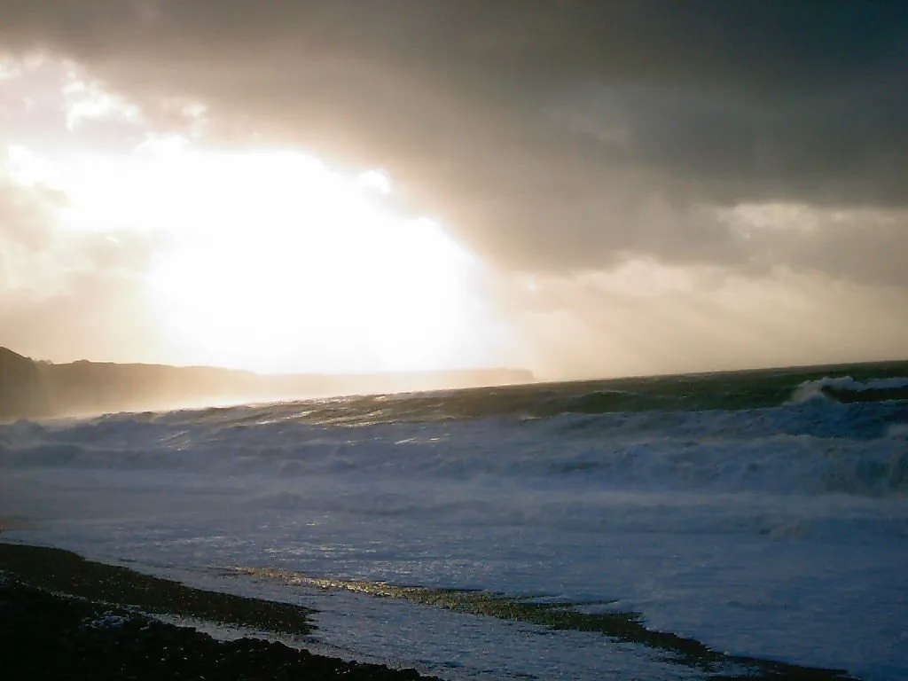 Waves crash onto a dark, pebbly beach under a dramatic, cloudy sky with the sun breaking through.