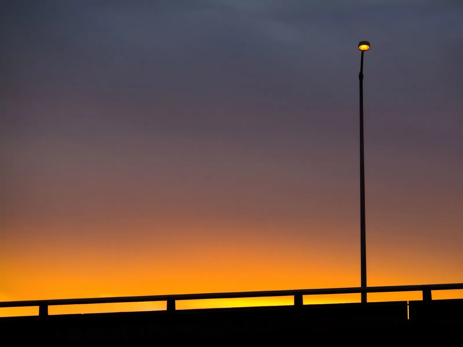 A single streetlight glows orange against a gradient sky of purple and orange, silhouetting a bridge railing.