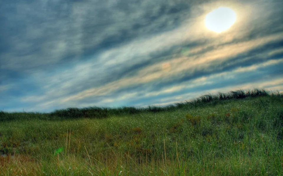 The sun shines brightly through a cloudy sky above a rolling dune covered in green grass.