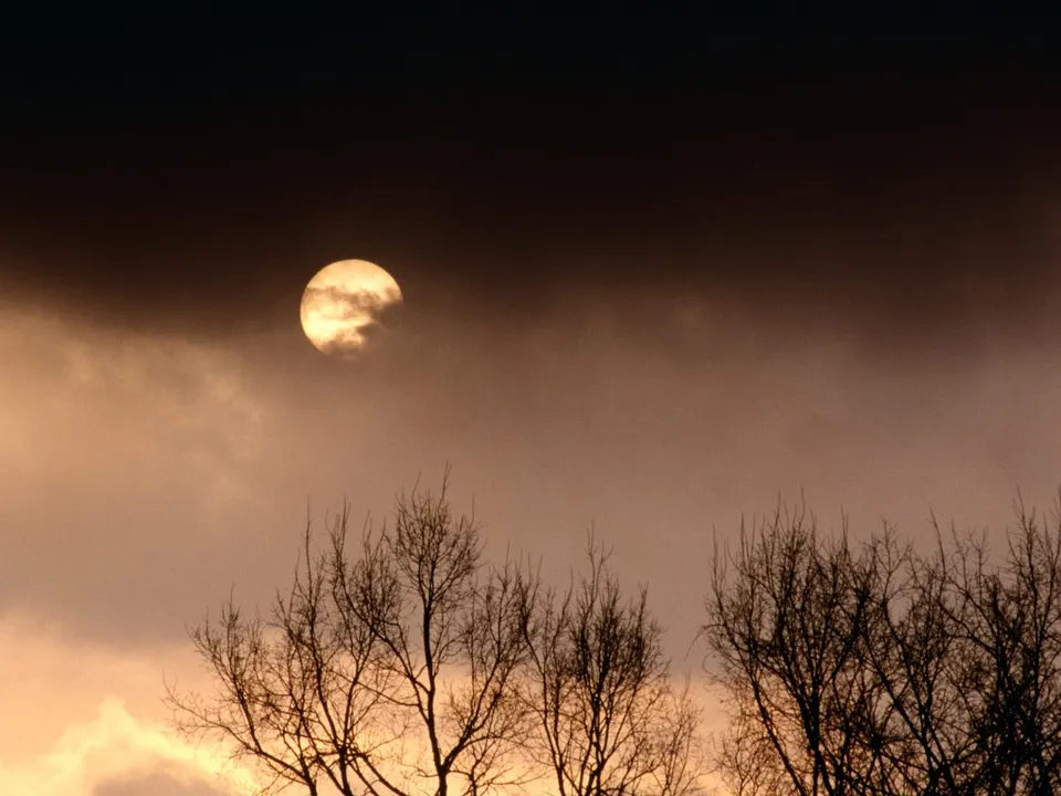 The sun is partially obscured by clouds, casting a warm glow against a dark, moody sky with silhouetted trees in the foreground.