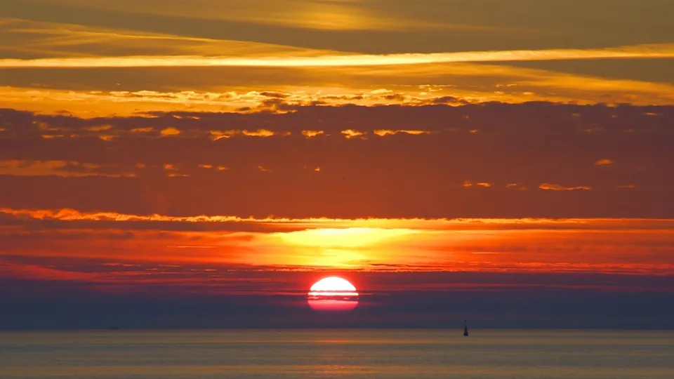 The sun is setting over a vast ocean, touching the horizon with dramatic clouds.