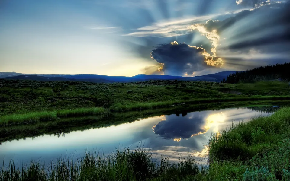 Sunbeams radiate from behind a dark cloud over a calm river reflecting the sky and landscape.