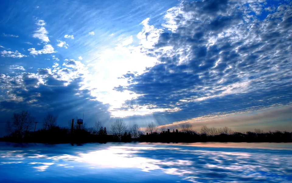 Sunbeams radiate through dark clouds, illuminating a reflective body of water with a silhouetted treeline.