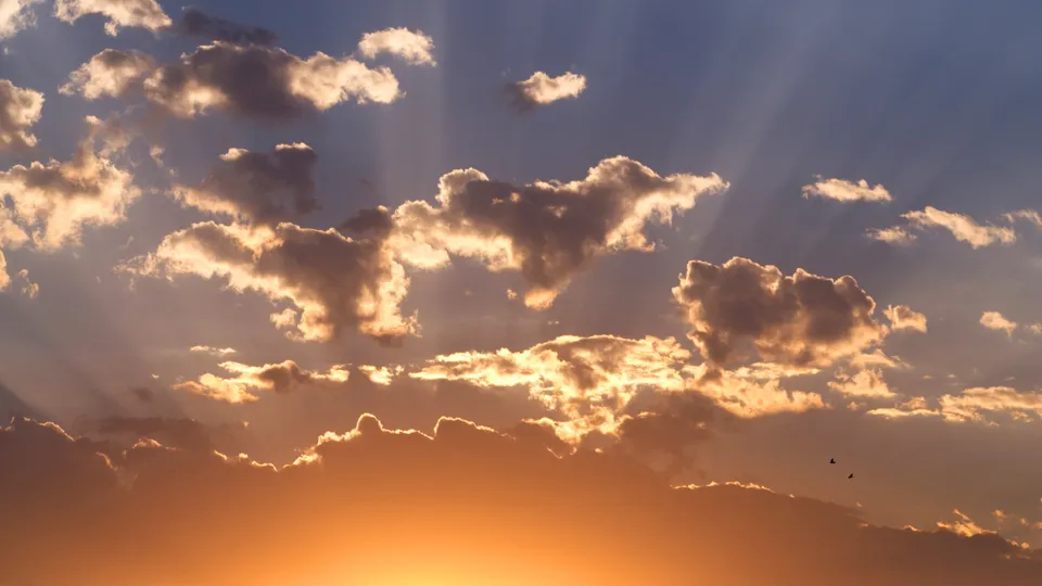 Sunbeams radiate through scattered clouds during a sunset, casting warm light.