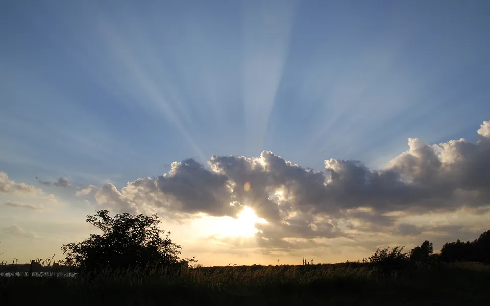 Sunlight streams through dark clouds at sunset, creating visible rays over a silhouetted landscape.