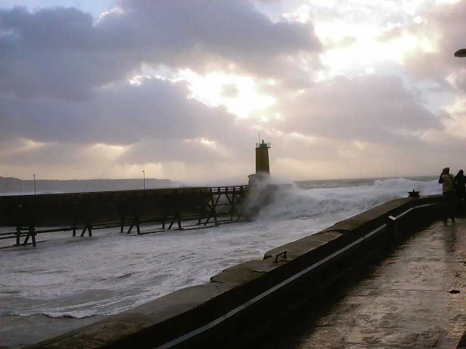 Sunbeams shine through clouds onto a lighthouse and a pier with crashing waves.