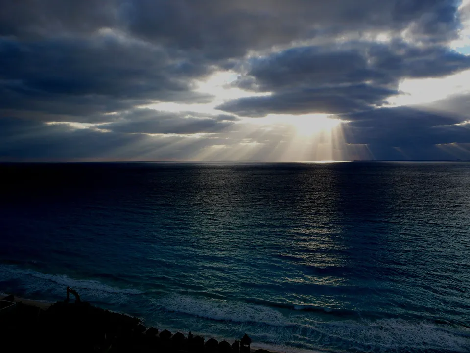 Sunbeams pierce through dark, dramatic clouds over a deep blue ocean with waves crashing on the shore.