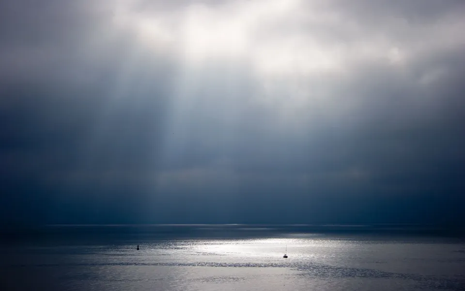 Sunbeams pierce through dark, dramatic clouds, illuminating a calm sea with a small sailboat.