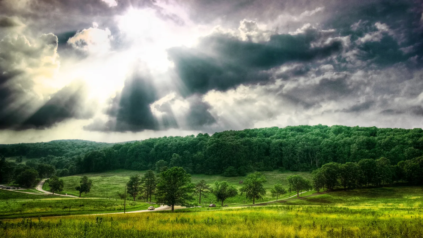 Sunbeams break through dark, dramatic clouds over a green, rolling landscape with trees.
