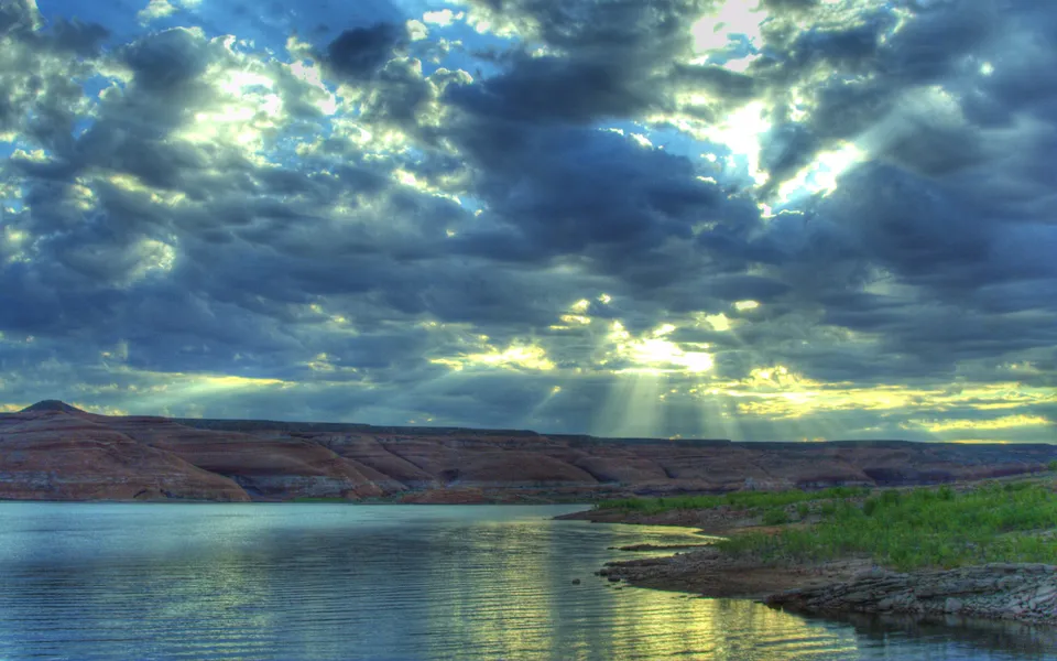 Sunlight streams through dark, stormy clouds over a lake, illuminating the water and distant hills.