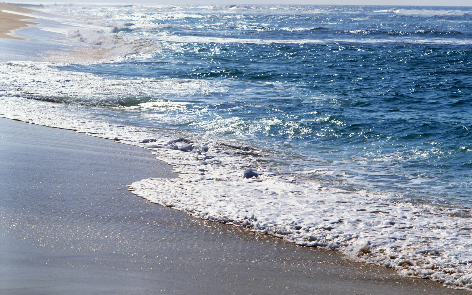 Deep blue ocean water sparkles with sunlight as gentle waves create white foam along the shore.